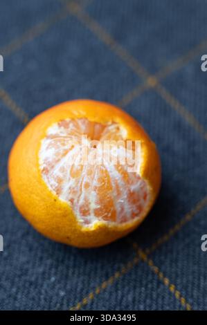 Halbgeschälte Mandarine auf kariertem Stoff, Ansicht von oben, betont Form und Farbe, klares natürliches Licht, saisonale Zutaten für Nachtisch oder Snack, Invit Stockfoto