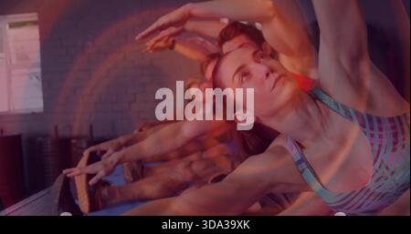 Frau in gemustertem Sport-BH, der den Arm im Studio auf blauen Matten hochstreckt, gestapelte Ausrüstung Stockfoto