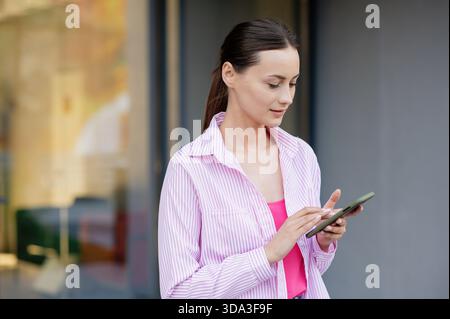 Porträt einer jungen brünetten Frau in rosa Hemd mit Smartphone im Freien Stockfoto