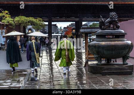 Mönche in grünen und anderen zeremoniellen Gewändern gehen im Regen durch das große Sanmon-Tor des Zenkoji-Tempels in Nagano; der bronzene Weihrauchbrenner ist im Vordergrund rechts zu sehen. Durch das Tor können Sie die Geschäfte auf Omotesando, dem historischen Pilgerweg zum Tempel, sehen. Nagano, Präfektur Nagano, Japan Stockfoto