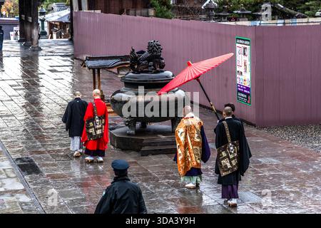 Buddhistische Mönche in zeremoniellen Gewändern (rot und Gold) gehen im Regen am großen bronzenen Weihrauchbrenner (香炉, Kōro) im Außenbereich des Zenkoji-Tempels in Nagano vorbei; ein stellvertretender Priester beschützt den Hohenpriester mit einem roten Wagasa-Ölpapierschirm. Der Weihrauchbrenner mit seinem Löwendeckel ist ein typisches Merkmal größerer Tempelkomplexe. Nagano, Präfektur Nagano, Japan Stockfoto