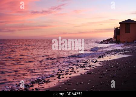 Pastellfarbener Sonnenuntergang über dem Mittelmeer an der spanischen Küste mit Wellen und Kiesstrand im Vordergrund. Stockfoto