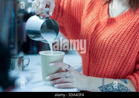 Horizontaler Blick auf den Barista, der weißen Milchstrom in die Tasse gießt, gehalten von tätowierter Hand in gemütlicher Café-Atmosphäre. Stockfoto