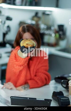 Weiblich in orangefarbenem Oberteil mit Handschuhen verlängert knuspriges Croissant mit Streuseln nach vorne in unscharfem Café-Hintergrund. Stockfoto
