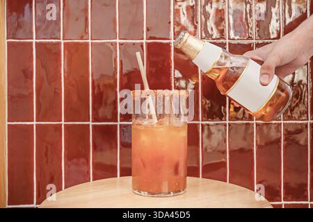 Eine Hand, die eine Bierflasche über einer braunen mexikanischen Michelada hält, mit einem Strohhalm auf einem Holztisch Stockfoto