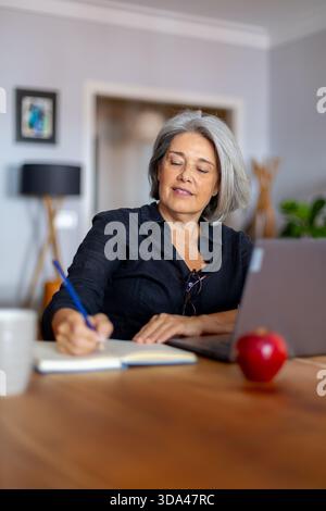 Reife Geschäftsfrau schreibt Notizen in Notizbuch, während sie von zu Hause aus mit einem Laptop arbeitet, mit einem Apfel auf dem Tisch Stockfoto