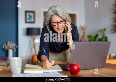Lächelnde, reife Geschäftsfrau, die ein Telefongespräch führt, während sie Notizen schreibt und effizient von ihrem Home Office aus arbeitet Stockfoto