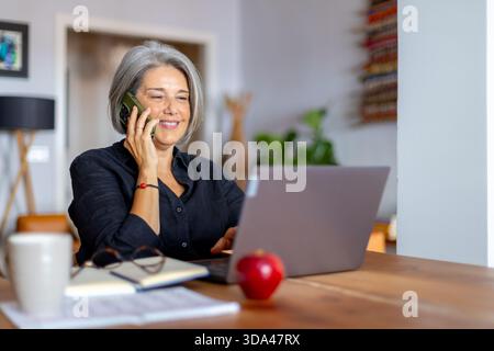 Fröhliche, reife Frau, die in gemütlicher Atmosphäre im Heimbüro unterwegs ist, während sie mit ihrem Smartphone chatten und auf ihrem Laptop tippen Stockfoto