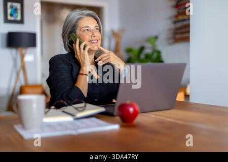 Lächelnde, reife Geschäftsfrau, die auf dem Mobiltelefon spricht und wegblickt, während sie zu Hause aus der Ferne arbeitet Stockfoto
