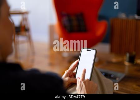 Frau, die sich auf einem bequemen Sessel entspannt, im Internet auf dem Handy mit leerem weißen Bildschirm, Mockup, über die Schulter aus der Nähe Stockfoto