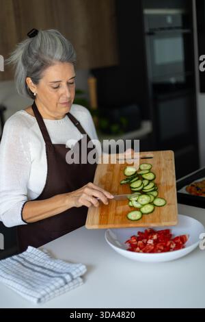 Frau, die vorsichtig geschnittene Gurken von einem Schneidebrett auf eine Schüssel mit gehackten Tomaten überführt, um eine nahrhafte Mahlzeit zu machen Stockfoto