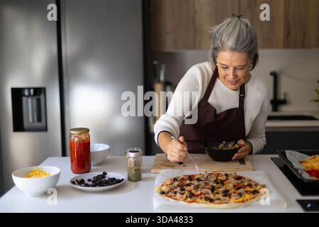 Seniorin trägt eine Schürze und bereitet eine köstliche hausgemachte Pizza für eine gesunde Mahlzeit in einer hellen Küche vor Stockfoto