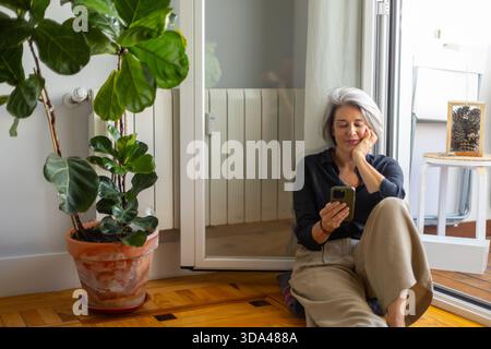 Entspannte, reife Frau, die auf dem Boden sitzt, mit dem Smartphone neben der Feigenpflanze im Topf, genießt einen ruhigen Moment zu Hause Stockfoto