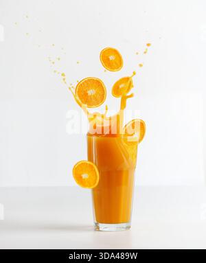 Glas mit Spritzern von Orangensaft und fallenden Orangenscheiben auf Tisch auf weißem Hintergrund. Gesundes Erfrischungsgetränk. Flüssige Bewegung. Vorderansicht. Stockfoto