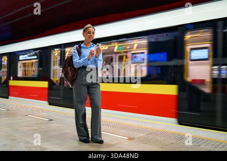 Lächelnde junge Frau mit Smartphone an einer U-Bahn-Station, die vor einem offenen Zug steht. Das Konzept des Pendelns, des städtischen Lebens, der Geschäftsreise, des modernen Lebens Stockfoto
