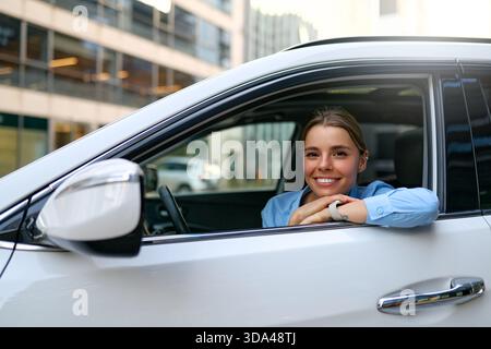 Lächelnde Geschäftsfrau in blauem Hemd, die ein Auto durch die Stadt fährt. Selbstbewusste Fahrerin, die die Fahrt genießt. Konzept der Mobilität, Unabhängigkeit, Modus Stockfoto