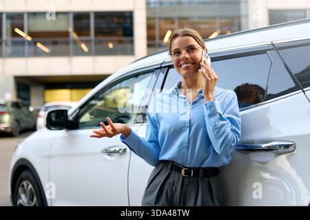 Glückliche Geschäftsfrau in blauem Hemd, die am Telefon plaudert, sich auf weißes Auto in städtischer Umgebung stützt. Professionelle Frau, die Gespräche genießt, modernes Stadtleben Stockfoto