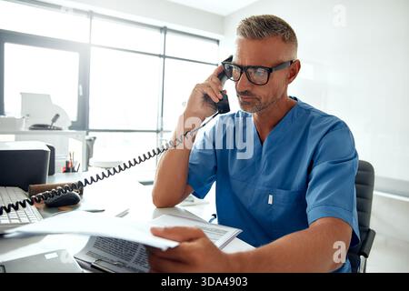In einer hellen und modernen Büroumgebung ist ein konzentrierter Arzt in Schutt-Kleidung am Telefon, führt Gespräche und dreht sich gleichzeitig Stockfoto