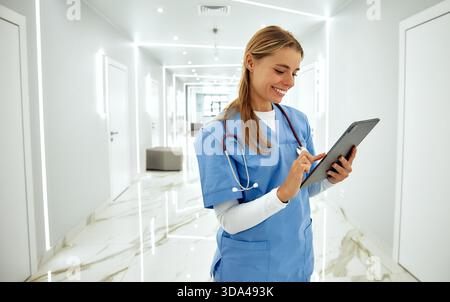 In einem hellen und modernen Krankenhausflur interagiert eine freundliche Krankenschwester mit blauen Peelings mit einer Tablette und demonstriert ihr Mitgefühl und p Stockfoto
