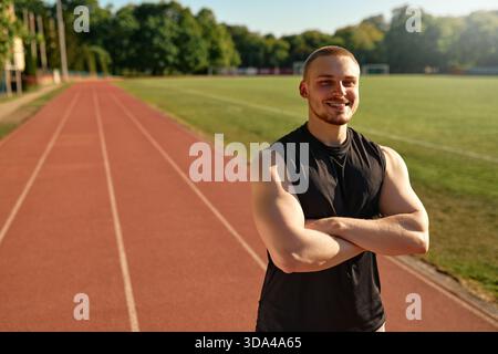 Glücklicher, sportlicher Mann, der mit gefalteten Armen auf der Laufbahn steht und selbstbewusst unter hellem Sonnenlicht lächelt. Konzept: Outdoor Fitness, Selbstwertgefühl, Position Stockfoto