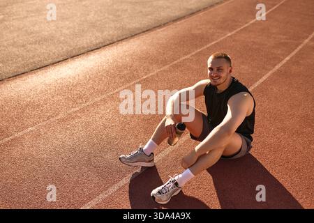 Junger muskulöser Mann in Sportbekleidung, der auf der Schiene sitzt und nach dem Training lächelt. Eine Wasserflasche halten und die Sonne genießen. Konzept: Hydratation, gesunde LIFE Stockfoto