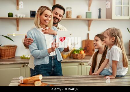 Eine Familie versammelt sich in der Küche, eine Frau liest eine Karte mit einem roten Herzen darauf. Sie lächelt, während ihr Mann und zwei junge Töchter aufmerksam zusehen Stockfoto