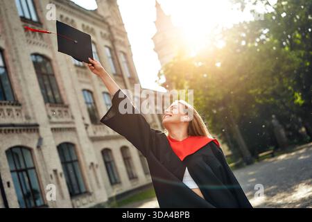Eine Absolventin feiert ihre akademische Leistung mit Freude, indem sie ihre Kappe auf einem renommierten Universitätscampus in die Luft wirft und damit Erfolg A symbolisiert Stockfoto