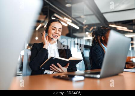 Team erfolgreicher Callcenter-Mitarbeiter mit Headsets, die mit Laptops am Tisch sitzen und Anrufe entgegennehmen und Kunden helfen und unterstützen Stockfoto