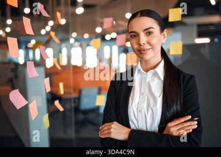 Junge Geschäftsfrau im formellen Anzug, die Haftnotizen mit Haftnotizen auf Glas klebt, erinnert an kreatives Brainstorming in einem modernen C Stockfoto