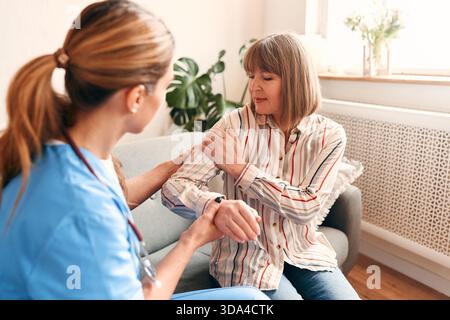 Ein Hausarzt zu Hause in medizinischer Uniform mit einem Stethoskop, der eine ältere Frau untersucht, die sich über Schmerzen im Arm beschwert, während sie auf dem Sofa sitzt Stockfoto