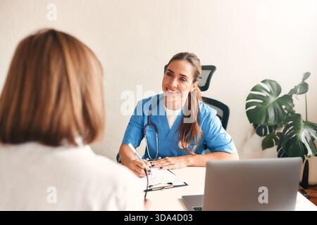 Ärztin in medizinischer Uniform mit Stethoskop am Tisch mit Laptop Notizen machen und mit dem Patienten in der Arztpraxis im Krankenhaus sprechen. Hea Stockfoto