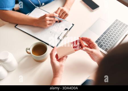 Ärztin in medizinischer Uniform mit Stethoskop am Tisch mit Laptop Notizen machen und mit Patienten über Pillen in der Pillbox sprechen. Gesundheitswesen Stockfoto
