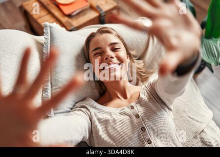Junge Frau, die auf einem Sofa in einem gemütlichen Wohnzimmer zu Hause liegt und sich an einem freien Tag entspannt, lächelt und zur Kamera greift. Stockfoto