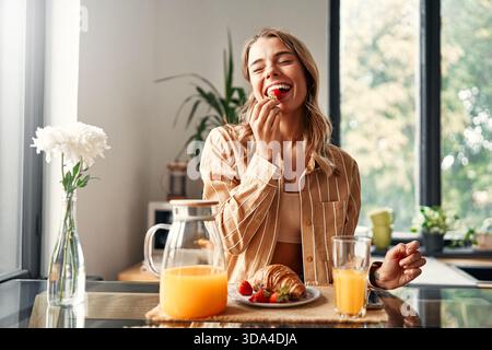 Junge glückliche Frau, die am Tisch in der Küche sitzt, Croissants mit Erdbeeren isst und Saft trinkt. Frau frühstückt in einer gemütlichen Küche Stockfoto