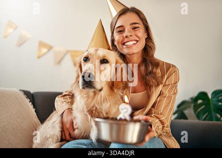 Junge glückliche Frau, die Geburtstagskuchen hält, den Geburtstag ihres Haustierhundes feiert, sich umarmt und Spaß mit ihrem Haustier hat, während sie im Wohnzimmer bei h sitzt Stockfoto