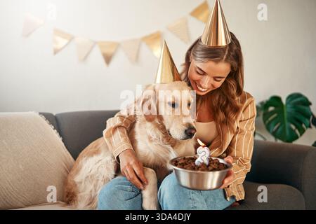 Junge glückliche Frau, die Geburtstagskuchen hält, den Geburtstag ihres Haustierhundes feiert, sich umarmt und Spaß mit ihrem Haustier hat, während sie im Wohnzimmer bei h sitzt Stockfoto