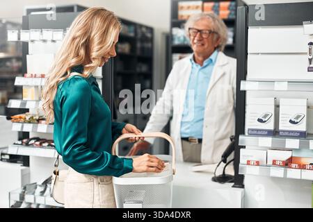 Ein leitender männlicher Apotheker in einem weißen Mantel hilft einer Kundin an einer modernen Apotheke. Sie lächeln und reden. Konzept: Kundendienstleistungen Stockfoto