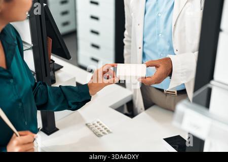 Ein älterer männlicher Apotheker im weißen Mantel hilft einer lächelnden blonden Frau an der Kasse in einer modernen Apotheke. Sie plaudern und lächeln. Konzept Stockfoto