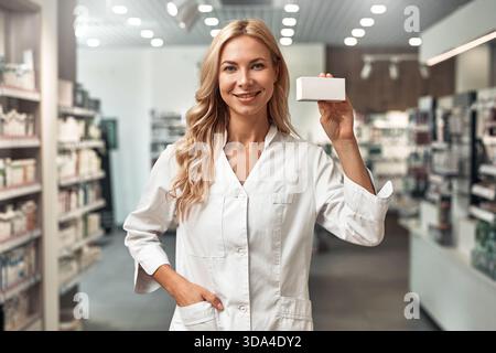 Selbstbewusste lächelnde Apothekerin in weißer Uniform, die eine leere Medizinschachtel hält, während sie in einem modernen Apothekeninnenraum steht. Medizinisches Fachpersonal Stockfoto