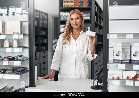 Blonde Apothekerin, die hinter der Theke in einer modernen Apotheke steht, eine leere weiße Medikamentenbox hält und selbstbewusst lächelt. Konzept: Gesundheit ca. Stockfoto