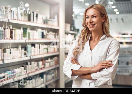 Blonde Apothekerin im weißen Mantel, die in einer modernen Apotheke steht und selbstbewusst lächelt. Professionelles Gesundheitskonzept. Stockfoto