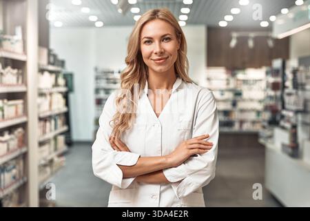Blonde Apothekerin im weißen Mantel, die in einer modernen Apotheke steht und selbstbewusst lächelt. Professionelles Gesundheitskonzept. Stockfoto