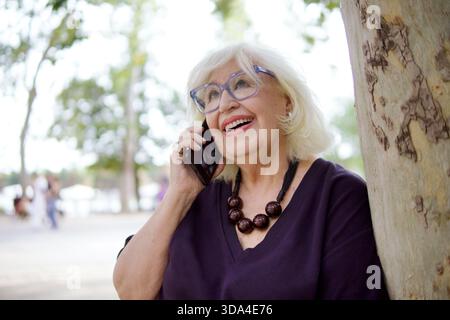 Ältere Frau lacht, während sie draußen telefoniert, mit Brille und einer stilvollen Halskette Stockfoto