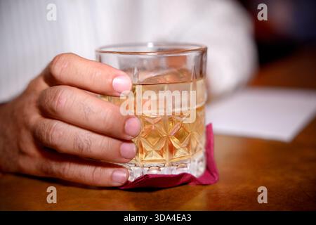 Hand hält ein Kristallglas Whiskey mit Eis auf einem Tisch, mit einem weißen Blatt Papier im Hintergrund Stockfoto