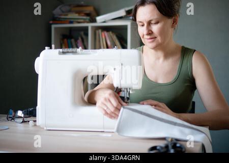 Nahaufnahme einer fröhlichen Schneiderin, die lächelt, während sie an ihrer Nähmaschine im Studio arbeitet und ihr Handwerk liebt. Stockfoto