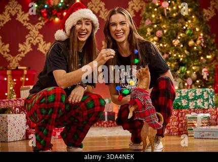 Zwei Schwestern und ihr Hund feiern Weihnachten im passenden karierten Pyjama mit verziertem Baum Stockfoto