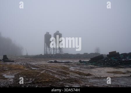 Zwei industrielle Lagersilos und ausgediente Reifen auf Silage oder Heu auf einem Bauernhof an sehr nassen und nebeligen Tagen Stockfoto