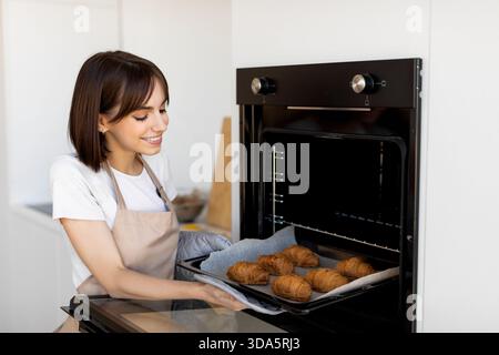 Die lächelnde Frau nimmt tagsüber frisch gebackene Croissants aus Oven in Modern Kitchen Stockfoto