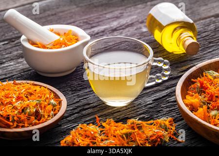 Calendula officinalis - Tasse Calendula-Tee, begleitet von getrockneten Blütenblättern und einem weißen Mörtel mit Calendula-Blüten Stockfoto