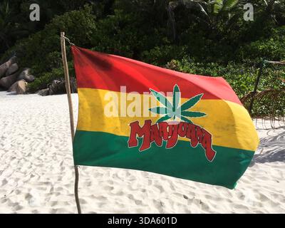 Flagge in jamaikanischen Farben mit einem großen Marihuanablatt und dem Wort „Marihuana“, das sanft im Wind winkt, am weißen Sandstrand mit grüner Küstenpflanze Stockfoto